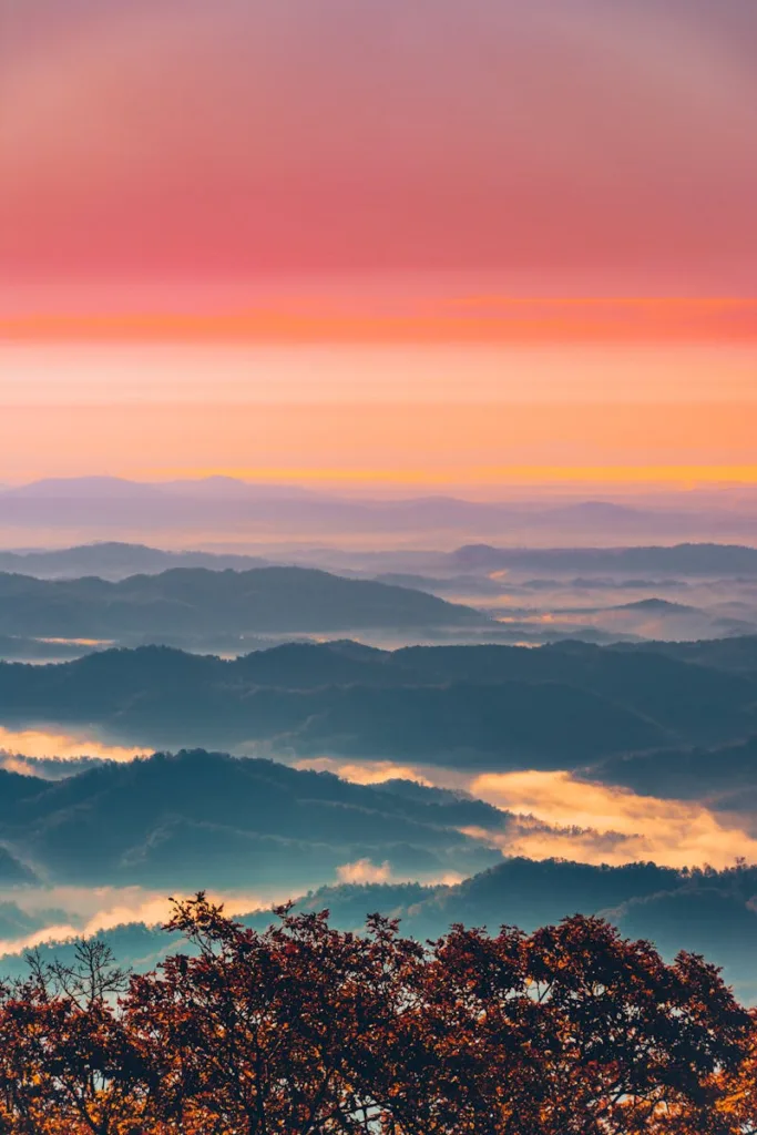 A breathtaking view of rolling mountains at sunset with fog and vibrant skies.